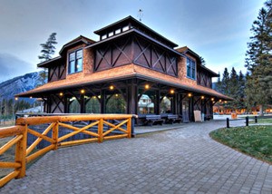 The Banff Park Museum, a rustic building with a wooden exterior, featuring prominent beams and a covered porch area. It appears to be located in a scenic, mountainous area, possibly a park or a historic site. In the background, there are tall trees and a glimpse of mountains. The foreground has a paved walkway and a wooden fence, and the overall atmosphere is tranquil and picturesque, likely captured during twilight, as lights are glowing under the porch.