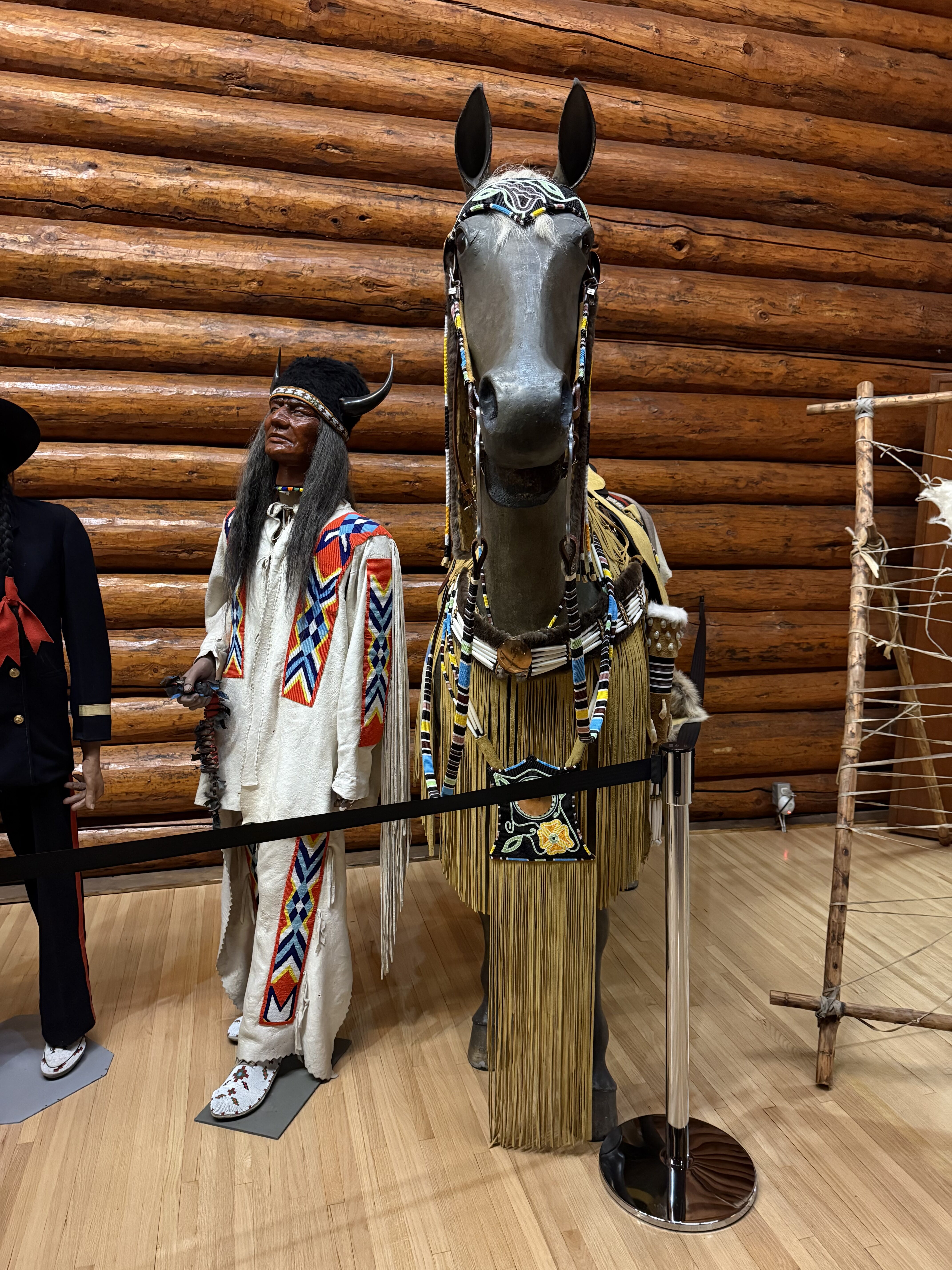 A display from the Buffalo Nations Museum. There is a mannequin dressed as a Native American figure standing next to a life-sized model of a horse. The mannequin is wearing traditional attire, including a long white garment with colorful geometric patterns in red, blue, and yellow. It also has a headdress with horns and long black hair. The horse model is adorned with decorative gear, including a headpiece and a blanket with similar colorful patterns and fringes. 