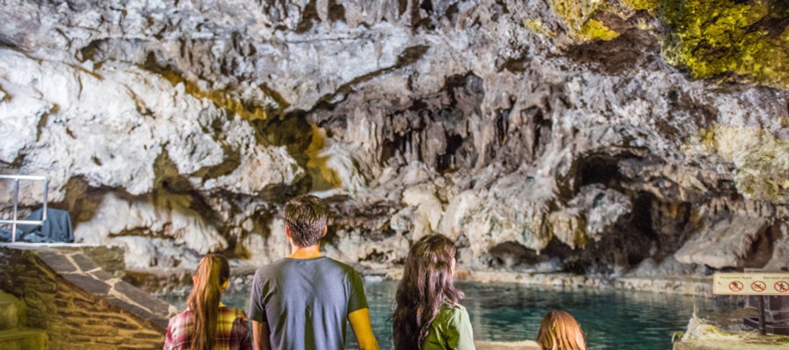 Four people facing away from the camera, looking at a large, illuminated cave or grotto interior. The rocky walls are rugged and feature a variety of textures. A pool of clear water sits at the base of the cave, reflecting the light. The group consists of two adults and two children. The atmosphere seems to be one of exploration and wonder as they observe the natural formations.