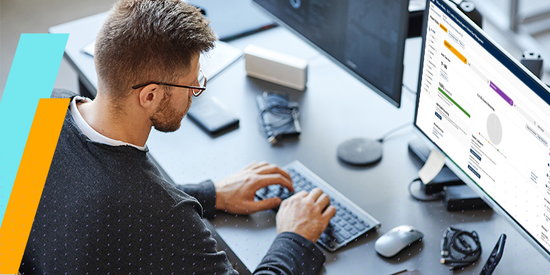 Man wearing glasses types on a keyboard at a desk with two large monitors displaying dashboards and data visuals.