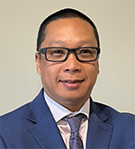 Professional headshot of a man in a blue suit, white shirt, and patterned tie wearing glasses.